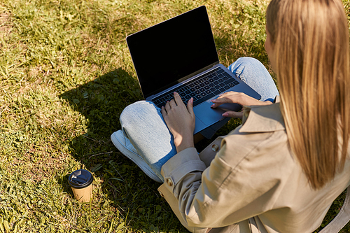 overhead view, woman in jeans and trench coat sitting on green lawn near paper cup and using laptop