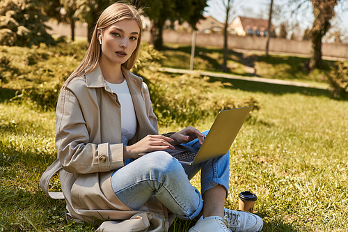 blonde woman in earphones and trench coat using laptop while sitting on grass near paper cup