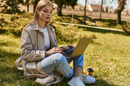blonde young woman in earphones and trench coat using laptop while sitting on grass near paper cup