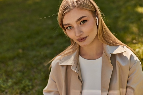 portrait of pretty young woman in wireless earphones and beige trench coat looking at camera in park