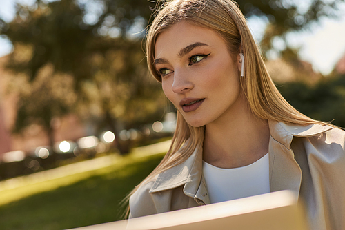 portrait of pretty young woman in wireless earphones and beige trench coat looking away in park