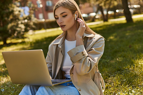 blonde young woman in wireless earphones and trench coat using laptop while sitting on grass