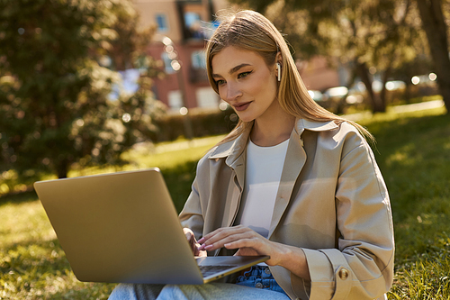 attractive young woman in wireless earphones and trench coat using laptop while sitting on grass