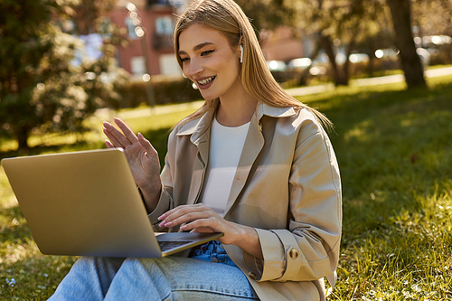 happy young woman in wireless earphones and trench coat using laptop while having video call