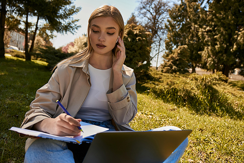 blonde woman in trench coat writing in notebook while using laptop and sitting on lawn in park