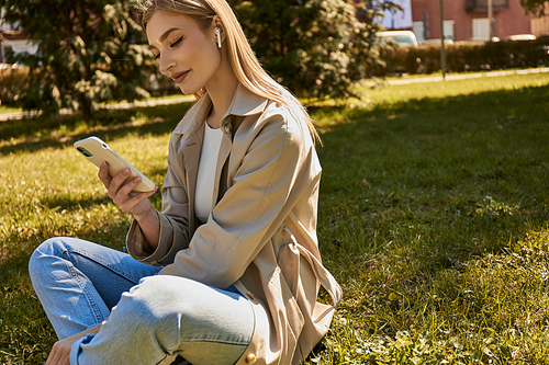 happy blonde woman in wireless earphones and trench coat using her smartphone and sitting on lawn