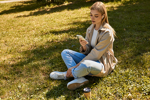 young blonde woman in wireless earphones and trench coat using her smartphone and sitting on lawn