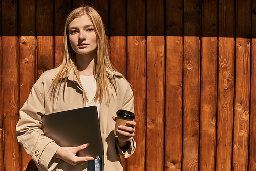 young blonde woman in stylish trench coat holding paper cup with coffee and laptop near fence