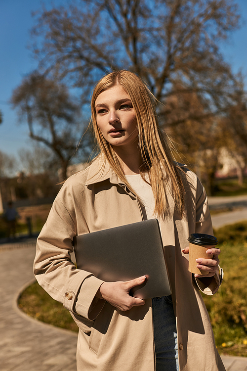young blonde woman in stylish trench coat holding paper cup with coffee and laptop in park
