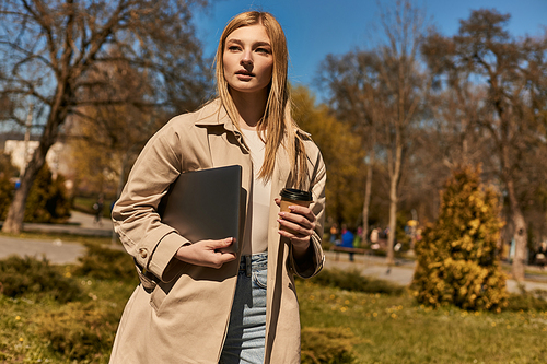 pretty blonde woman in stylish trench coat holding paper cup with coffee and laptop in park