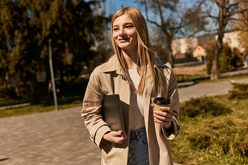 happy blonde woman in stylish trench coat holding paper cup with coffee and laptop in park