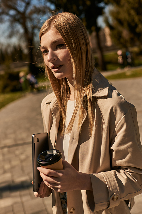 young blonde woman in beige trench coat holding paper cup with coffee and laptop and walking in park