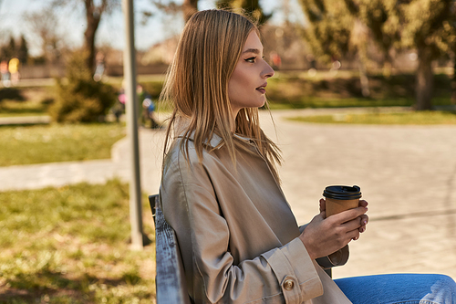 young blonde woman in trench coat holding paper cup with takeaway coffee, sitting on bench in park