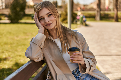 young happy woman in trench coat holding paper cup with takeaway coffee, sitting on bench in park