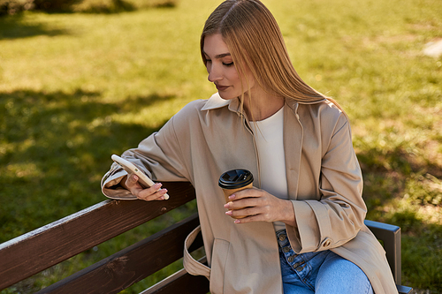 blonde woman in trench coat holding paper cup with coffee and using smartphone, sitting on bench