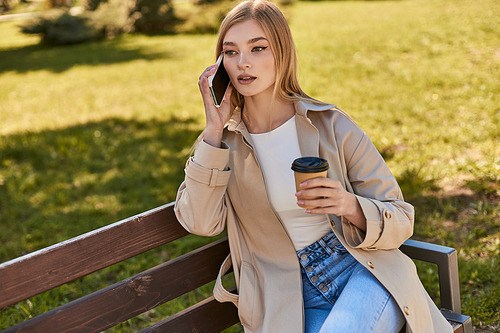 young woman in trench coat holding takeaway coffee and talking on smartphone while sitting on bench