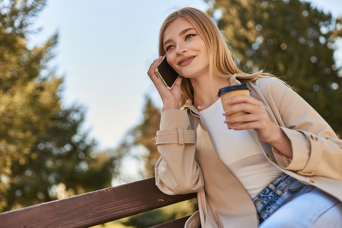 happy woman in trench coat holding takeaway coffee and talking on smartphone while sitting on bench