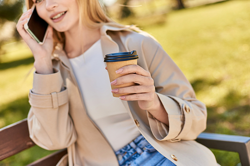 cropped woman in trench coat holding coffee to go and talking on smartphone while sitting on bench