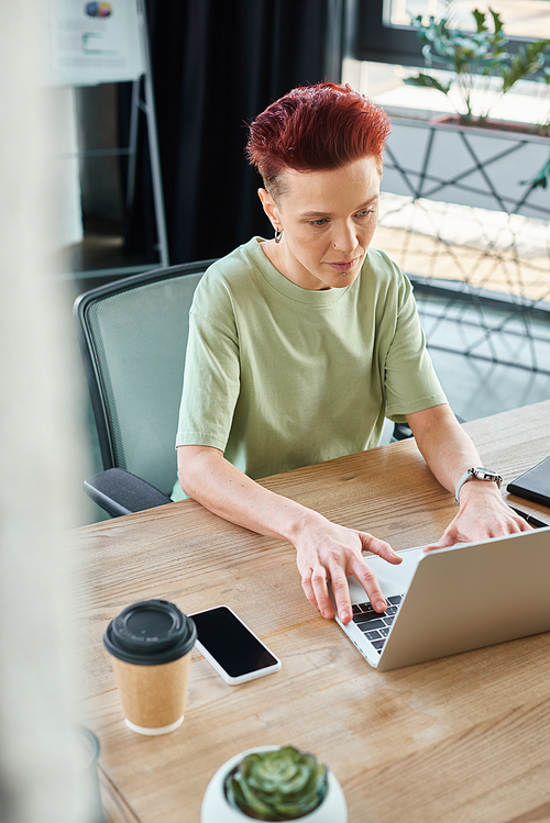 stylish ambitious queer person working at laptop near smartphone and coffee to go in modern office