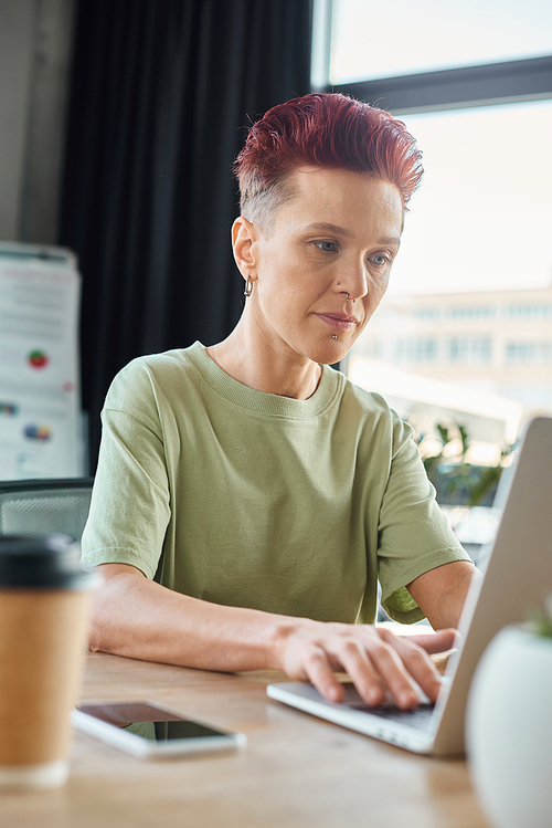 stylish queer person working at laptop near blurred smartphone and coffee to go in modern office