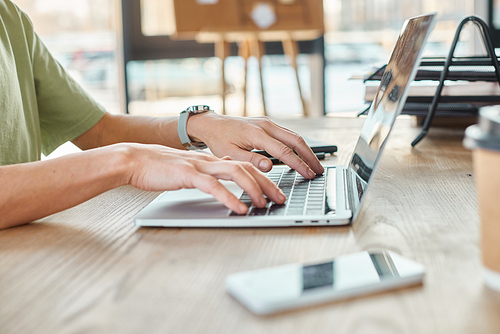 cropped view of queer person typing on laptop near blurred smartphone and coffee to go in office