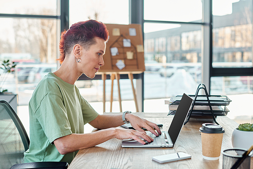 side view of non-binary person typing on laptop with blank screen near smartphone and coffee to go