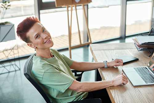 stylish happy non-binary person looking at camera white sitting near laptop at workplace in office