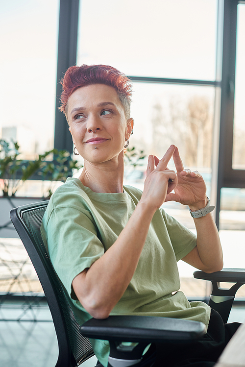 smiling short haired bigender person sitting and looking away at workplace in modern office