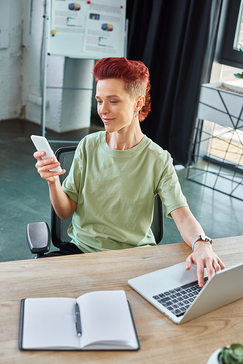 smiling bigender individual looking at smartphone near laptop and notebook in modern office