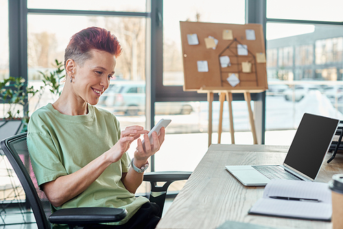 smiling bigender person using smartphone near laptop and notebook at workplace in modern office