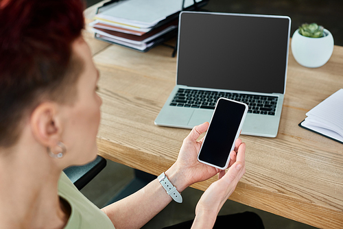 non-binary business person holding smartphone with blank screen near laptop at workplace in office