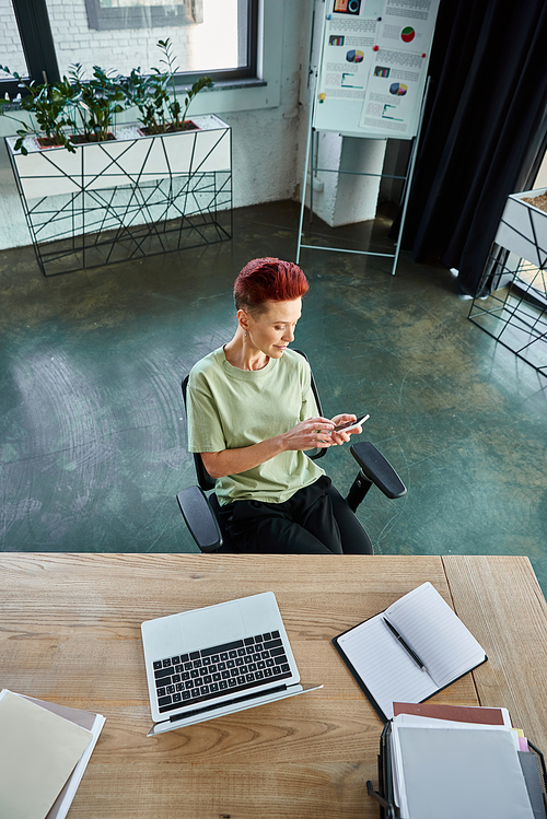 high angle view of queer manager messaging on smartphone near laptop and documents on work desk