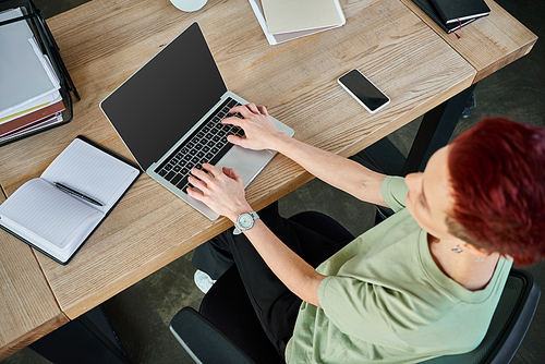 top view of queer person typing on laptop with blank screen near smartphone and notebook in office