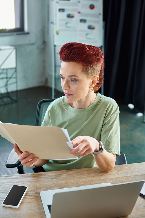 stylish queer manager working with documents near laptop and smartphone with blank screen in office