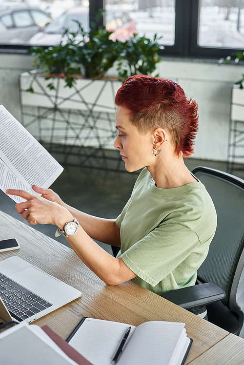 side view of stylish non-binary person working with documents near laptop and mobile phone in office