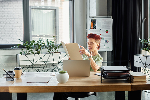 stylish bigender manager working with documents near laptop and coffee to go in paper cup in office