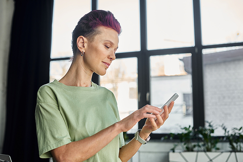 stylish queer manager in casual attire looking at smartphone while standing in modern office