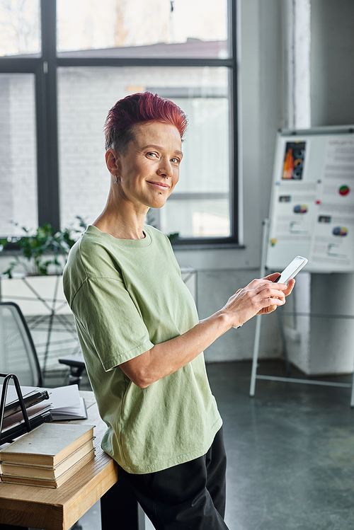 cheerful successful queer manager with mobile phone looking at camera while standing in office