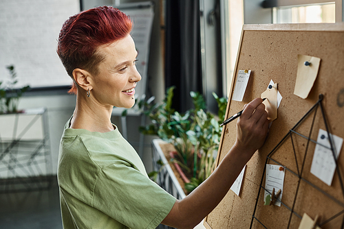 smiley bigender manager in casual attire writing on paper notes on corkboard in modern office