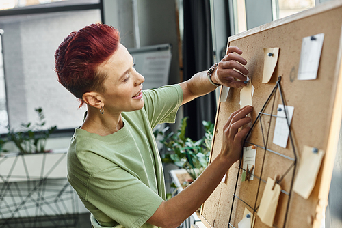 smiley bigender manager in casual attire pinning paper notes on corkboard in modern office