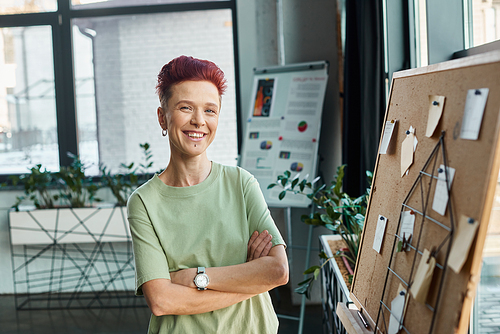 happy non-binary person with short hair looking at camera near corkboard with paper notes in office