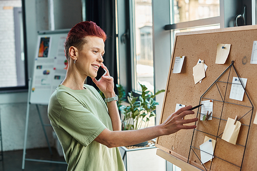 stylish queer person talking on mobile phone and looking at paper notes on corkboard in office