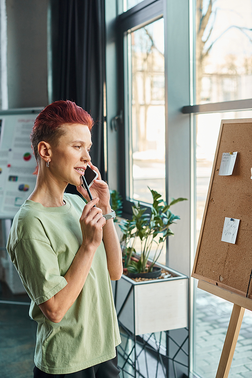 thoughtful queer person talking on mobile phone and looking at corkboard with paper notes in office