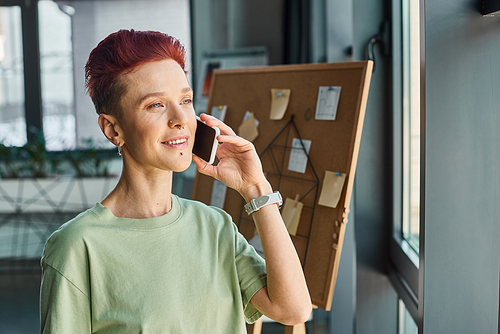 smiling bigender manager with short hair looking away during conversation on smartphone in office