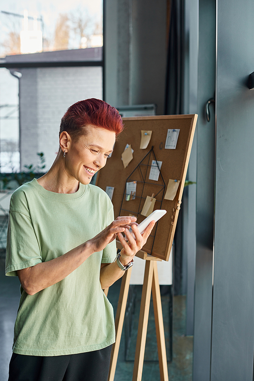 joyful short haired bigender manager in casual attire messaging on smartphone in contemporary office