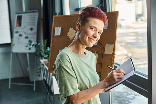 stylish queer manager smiling at camera and writing in notebooks in modern office environment