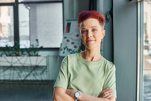 confident bigender business person with folded arms and looking at camera in office, portrait