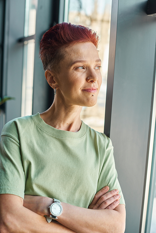 positive and dreamy bigender person with folded arms looking away through window in modern office