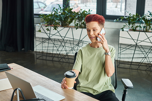 positive queer person with coffee to go in paper cup talking on mobile phone near laptop in office