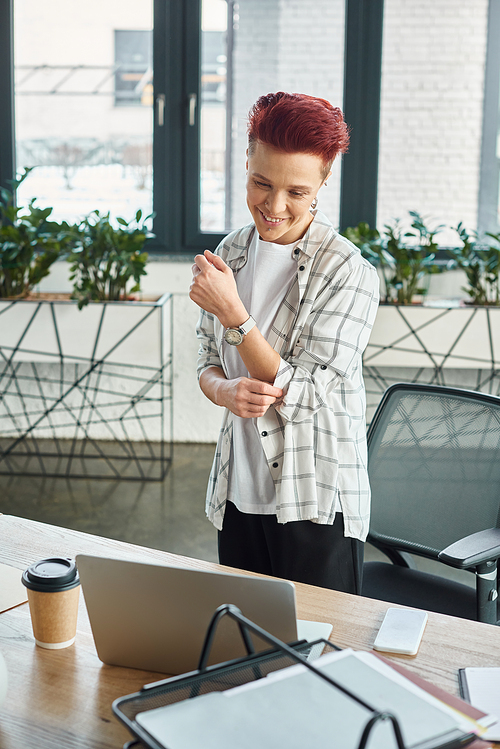 joyful bigender manager in casual attire standing at workplace and looking at laptop in office